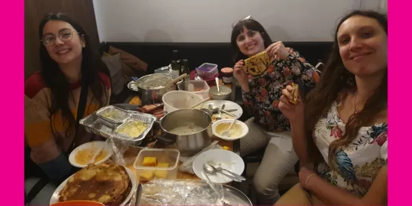 Three girls sitting at the table covered by the dishes brought by the participants. They smile and show some of the food.