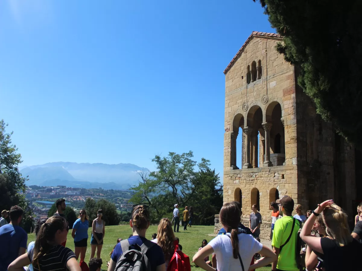 Several people standing in a circle in front of a romanesque church on top of a mountain.