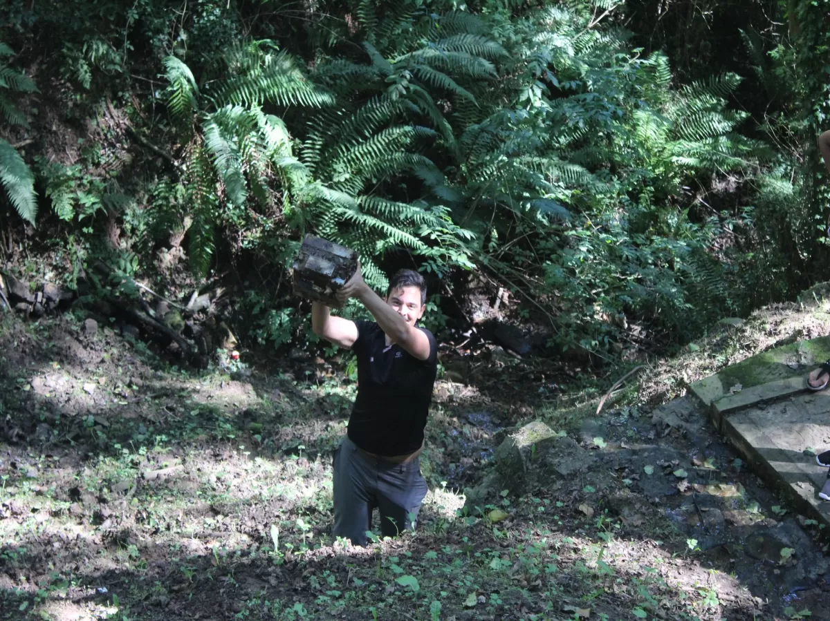 A very enthusiastic volunteer struggles to pick up a car battery in a forest.