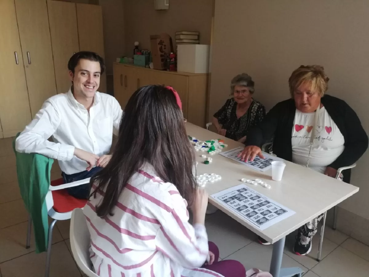 A handsome volunteer sits staring at the camera while three elderly women play games.