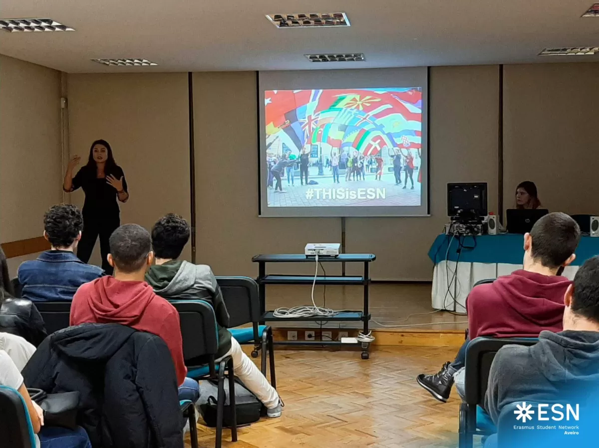 International student talking to a roomfull of local high school students. In the back, there is a screen showing a picture of people holding flags from many countries and the text "This is ESN".