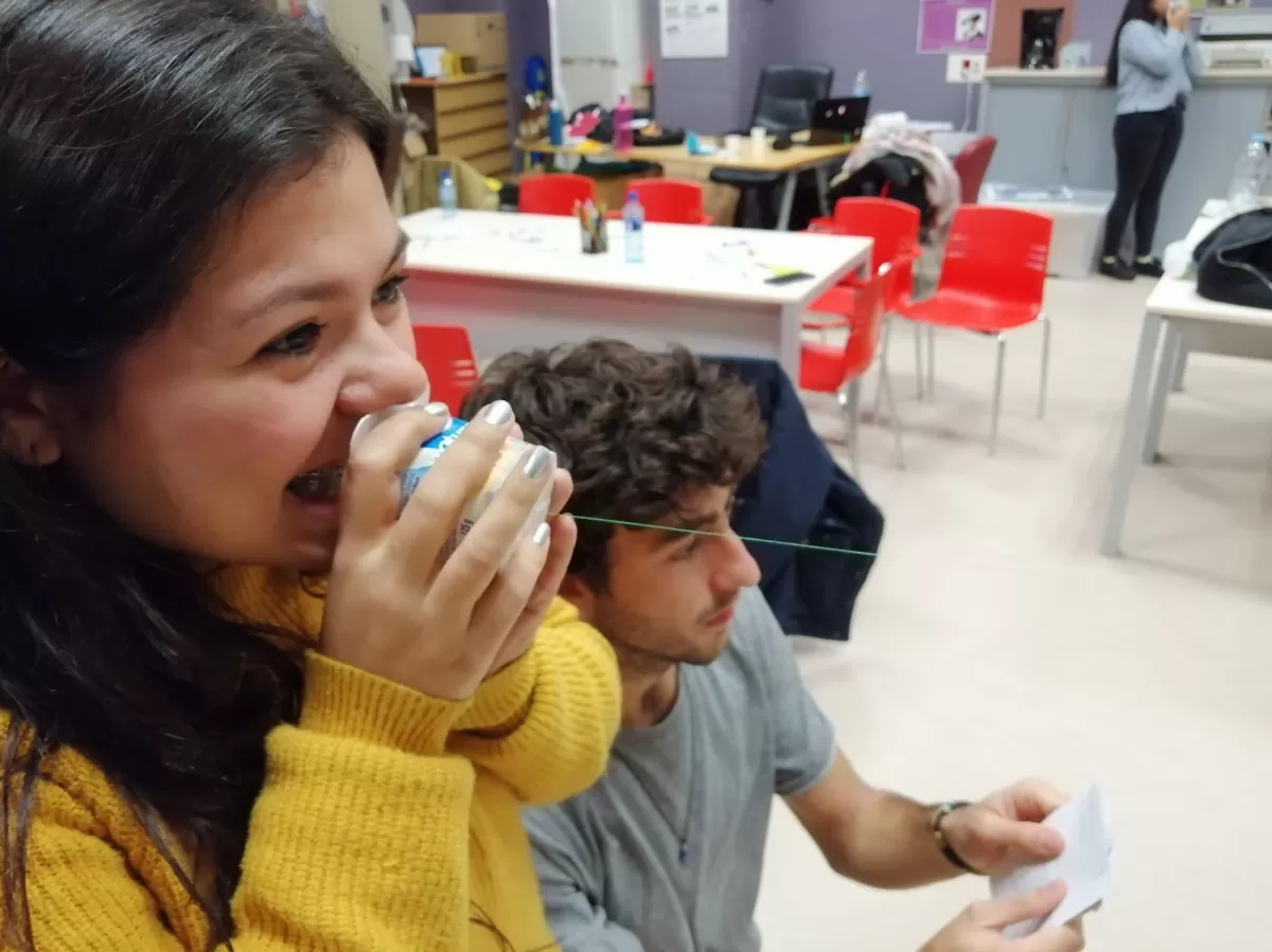 A girl talks to a yogurt cup tied to a string while a man reads something off a paper