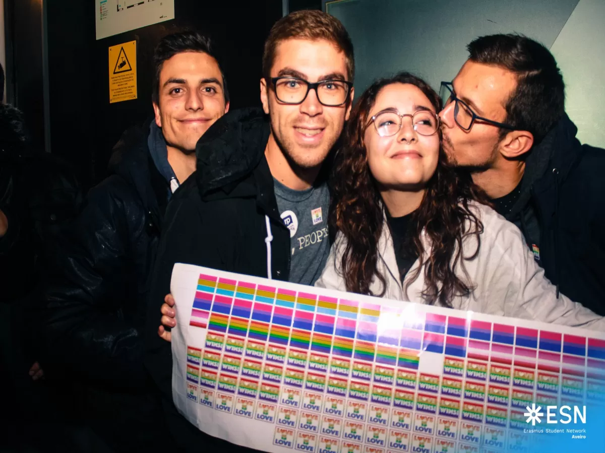 Four students smiling at the camera. One of them is holding a paper with stickers saying "Love is love" or with LGBTQ+ flags. 