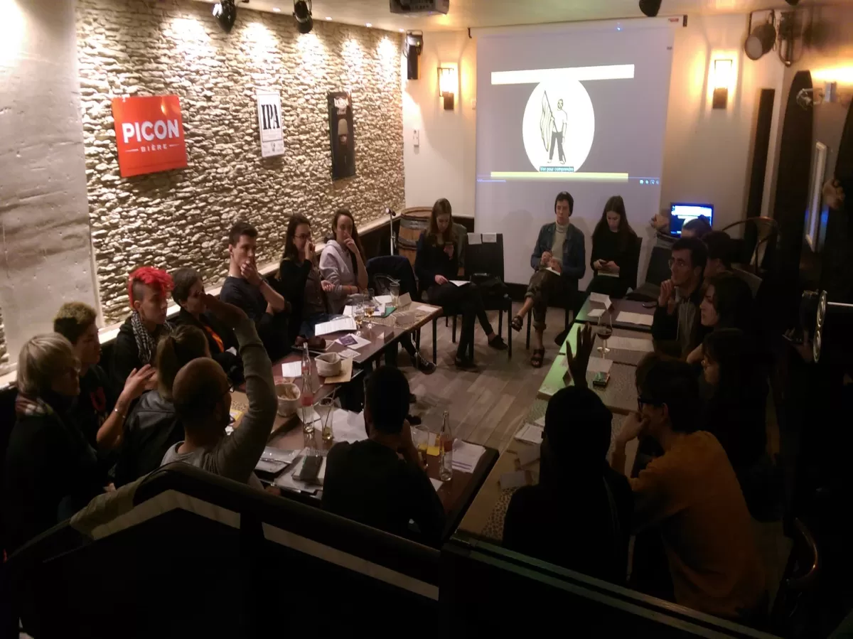 Picture of the going-on debate. they all face each others with tables in a  U form. Two persons are raising one hand to speak.  There is a screen in the background showing a drawing of a person holding a flag with stripes it come from the video we showed in the begining. 