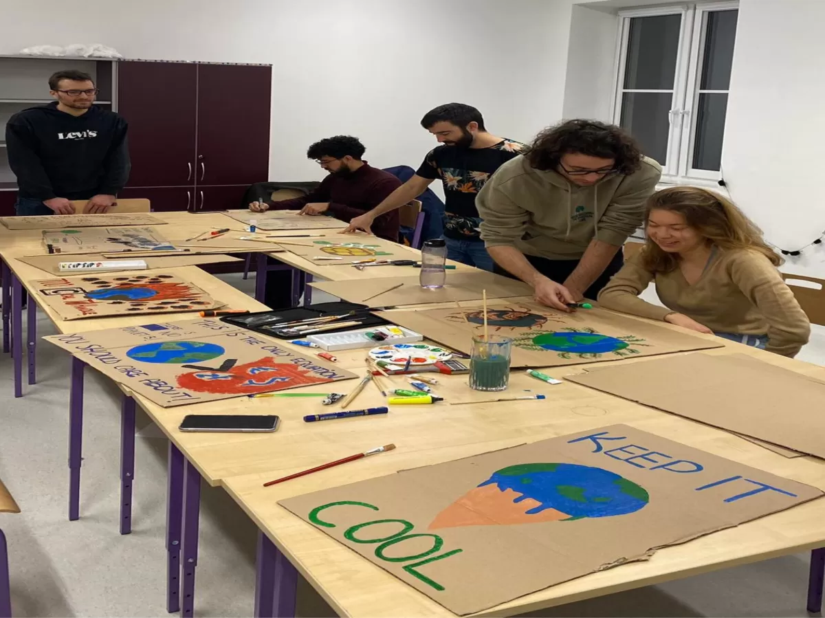 In the foreground there are banners, one of them says "keep it cool", in the background young people are gathered at a table and paint their banners.
