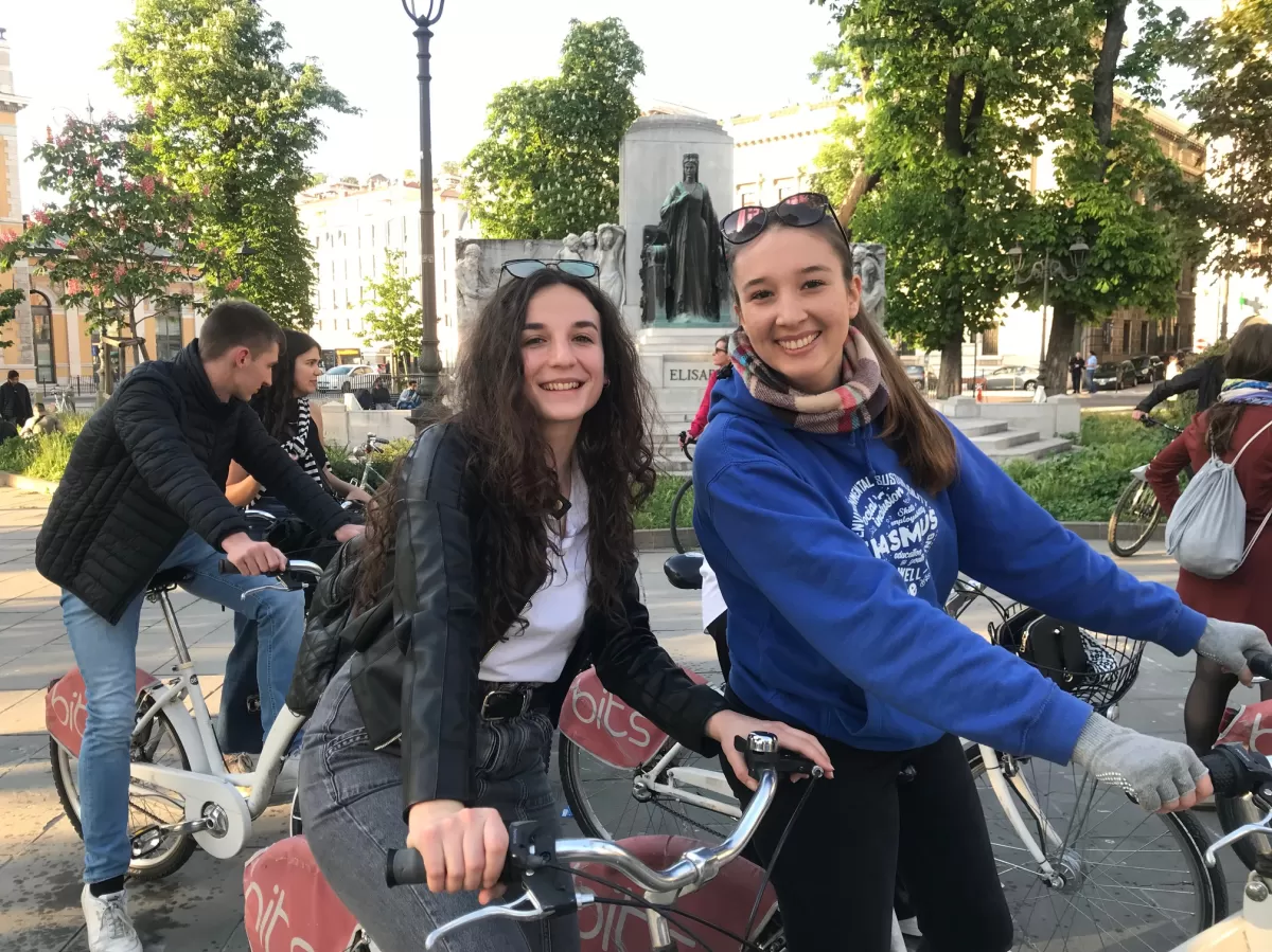 Two girls on their bikes are looking at the camera and smiling.  On the background there are more people, also on their bikes, talking to each other. Behind them there are some trees and a statue.