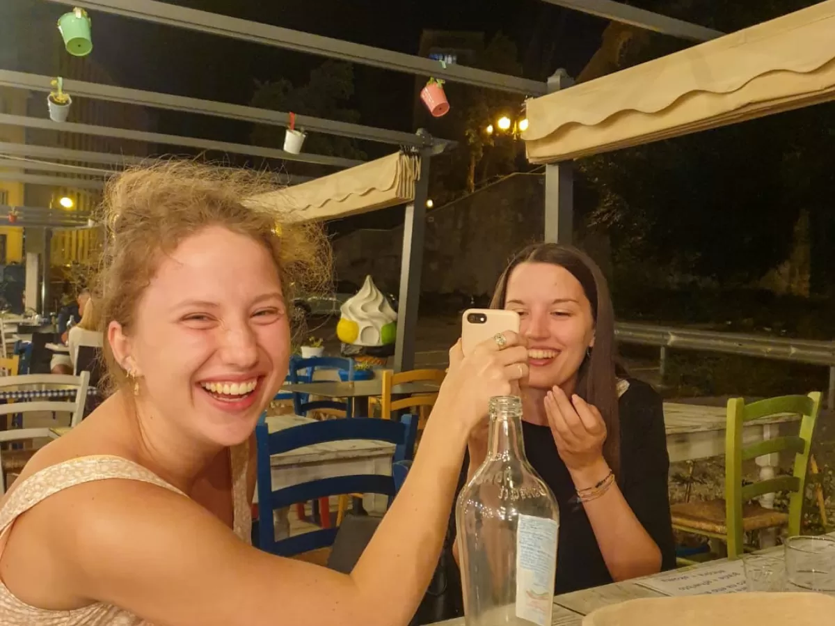 Two girls are sitting at a table. One of them is looking at the camera laughing and she is holding a phone in her hand, the other one is looking at the phone and she is also laughing.
