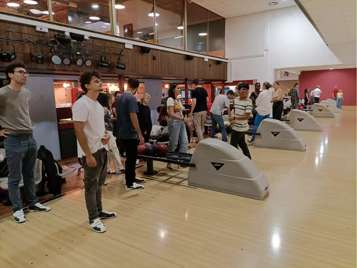The picture shows a group of people waiting for their turn to play bowling. Some of them are looking upwards (probably at the screen with the scores), some are laughing or talking to each other.