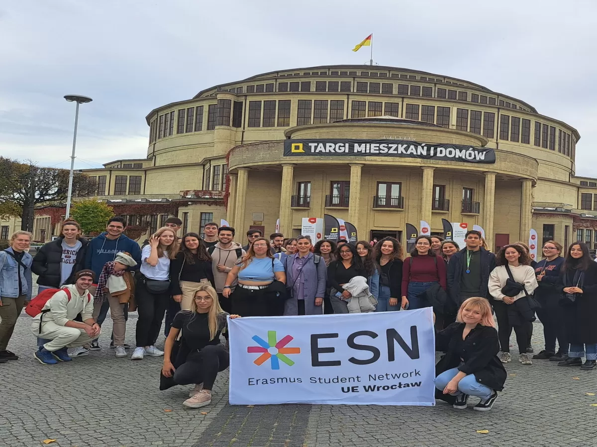 The photo shows a group of students posing for a group photo. In the background there is the building of the Centennial Hall. In the middle of the pictures two girls are holding a white ESN flag.
