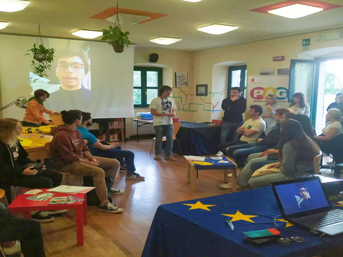 A group of people is sitting in a circle on some chairs and a man is standing between them, talking. Behind him we can see a screen where a video is being projected. In the foreground there is a table with a Europen flag lying on it and a computer on top of it.