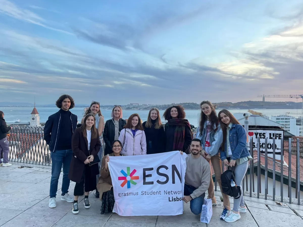 ESN members and Erasmus students holding up an ESN flag with a panoramic view of Lisbon's river behind