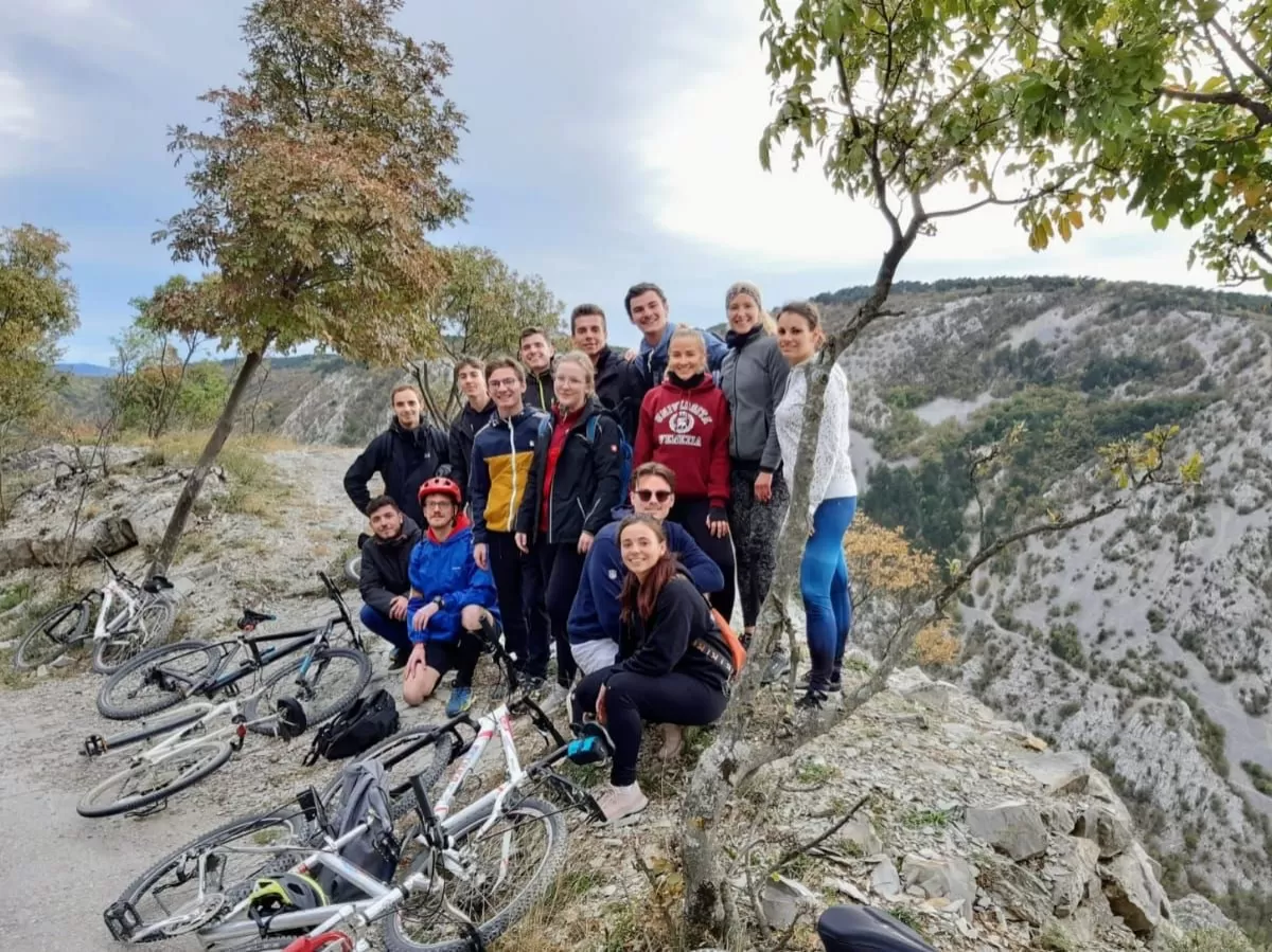 Group of people standing with rocks and trees behind them. Their bikes are laying on the ground in front of them. The picture is taken with a lateral perspective.
