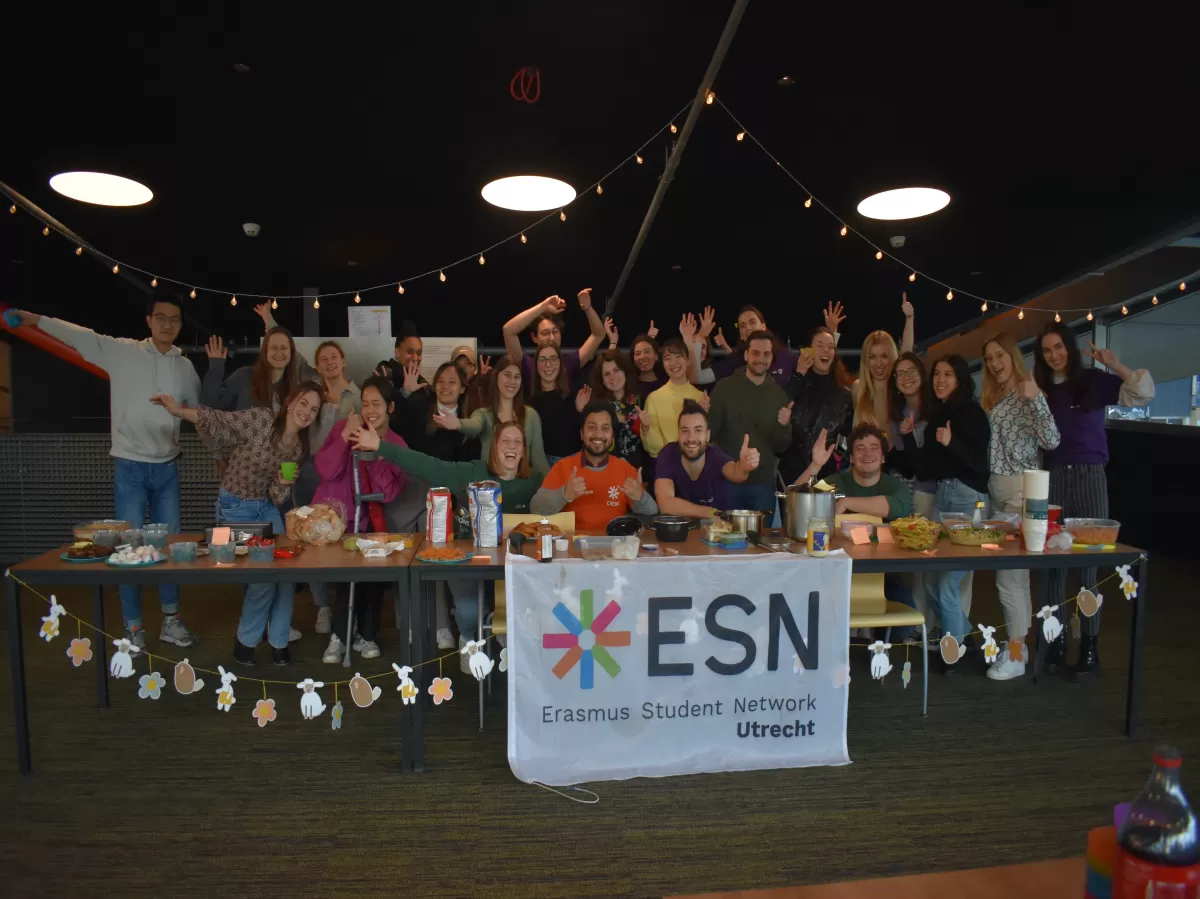 The image displays a group of people behind table full of different dishes. There is an ESN Utrech flag in the front.