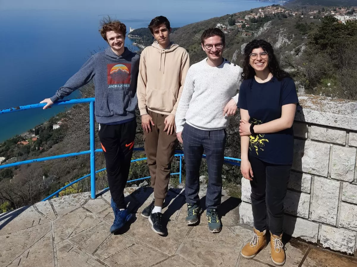 The picture shows 3 boys and a girl standing together, looking at the camera and smiling. Behind them is a view on the gulf of Trieste, with blue sea and sky, as well as the coast with trees and buildings..
