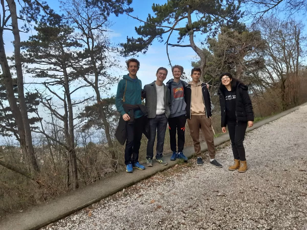 The picture shows 4 boys and a girl standing together in a group on a hiking trail, looking at the camera and smiling. There are some trees in the background, as well as a bright blue sky.