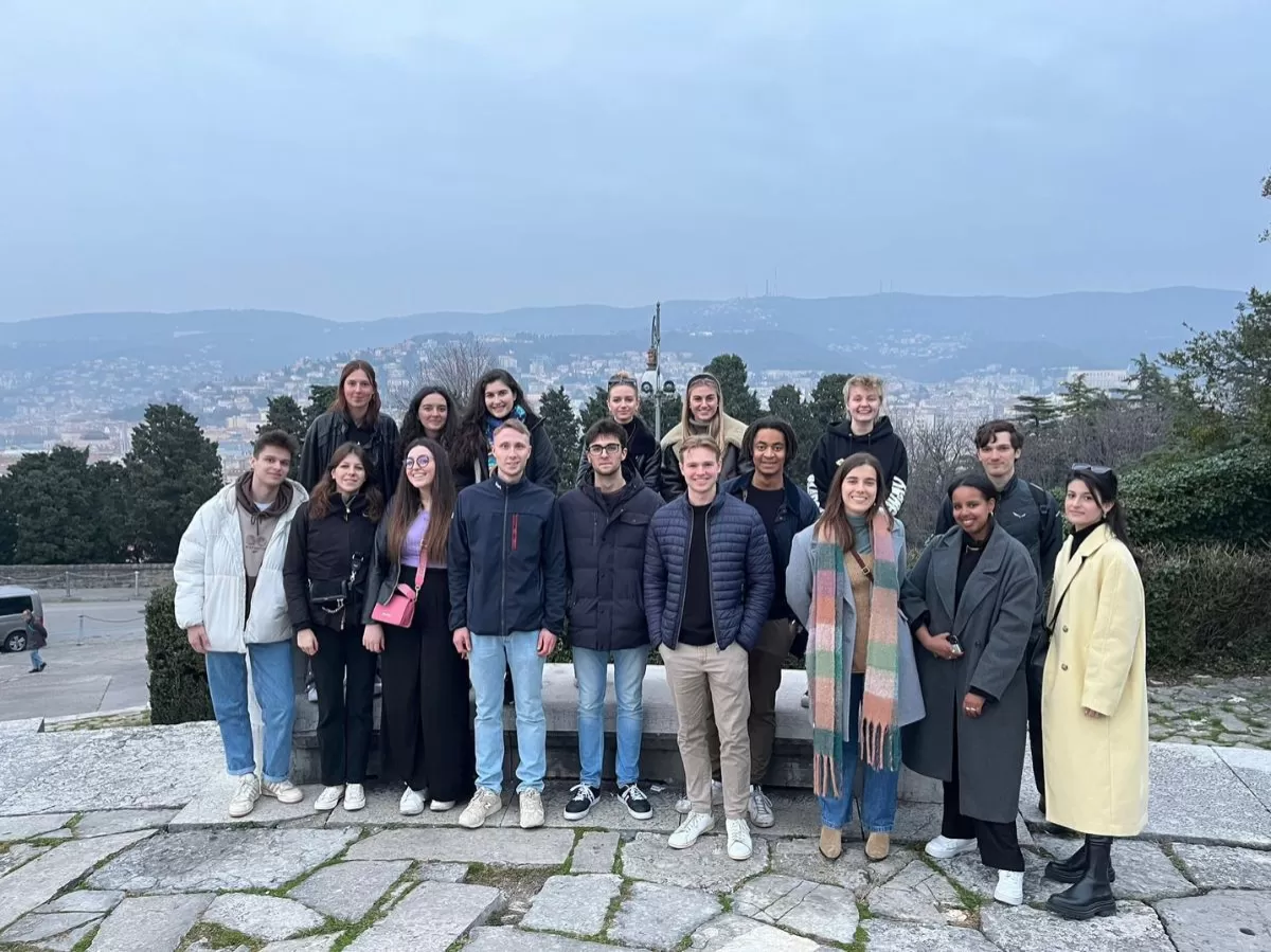 The picture shows the participants standing on the hill of San Giusto. Behind them there are some bushes, and on the background a view of the city of Trieste. The sky is cloudy.