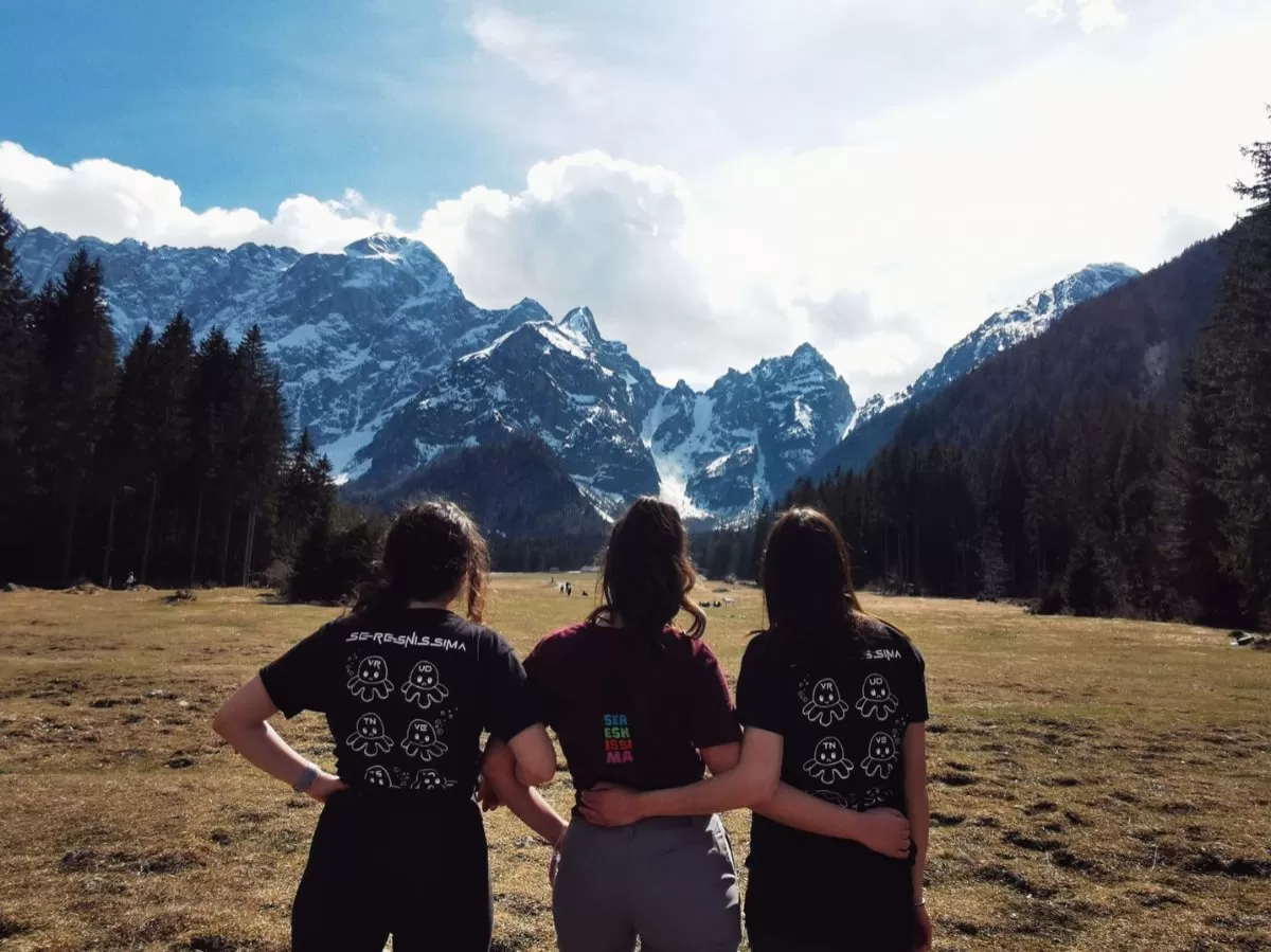 Three girls are standing with their backs at the camera, all of them are wearing the t-shirt of the SerESNissima Section Cooperation. In the background there are trees, snowy mountains and blue sky.