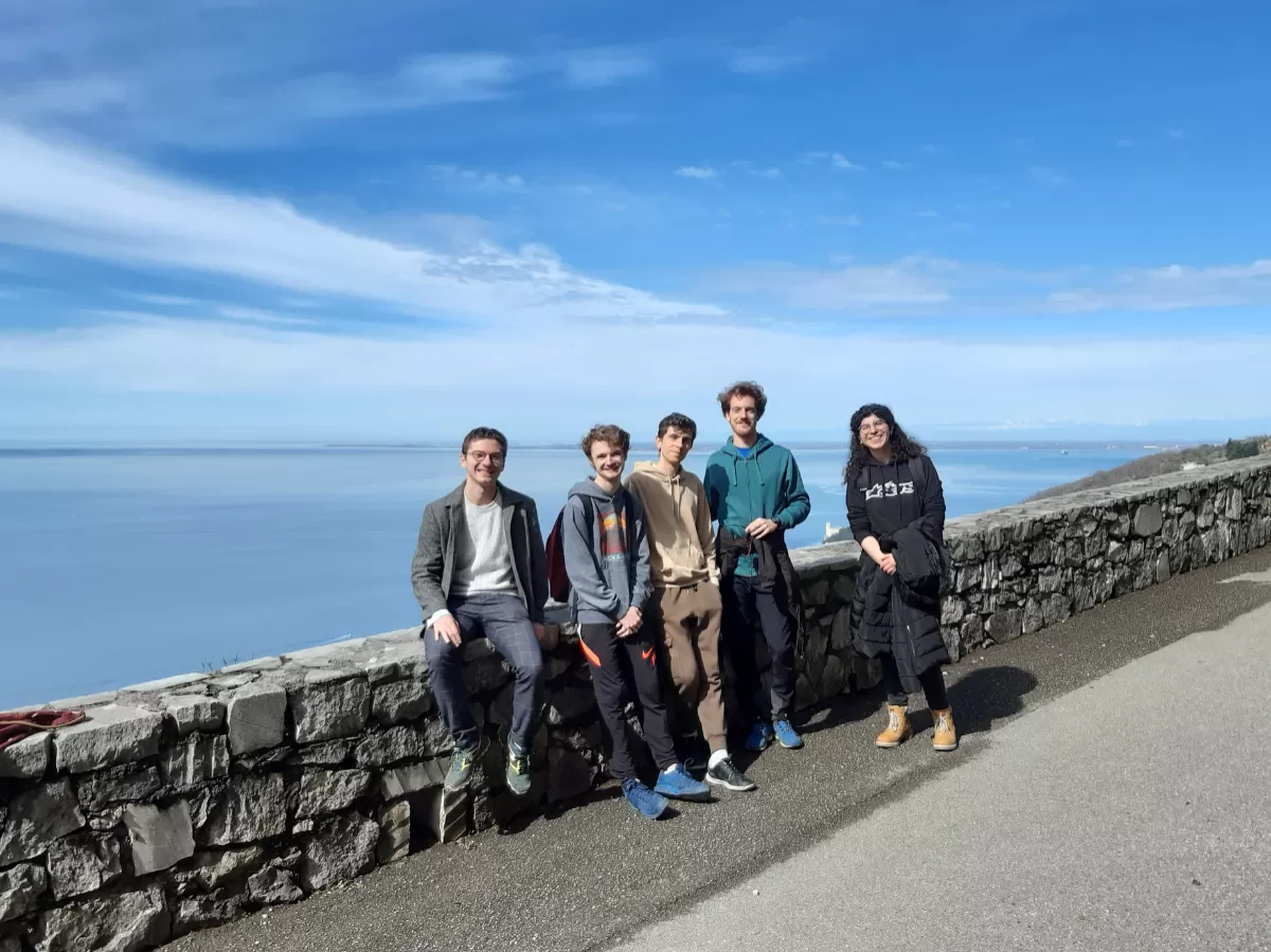 The picture shows 4 boys and a girl standing together in a group on a hiking trail, looking at the camera and smiling. There is the sea in the background, as well as a bright blue sky.