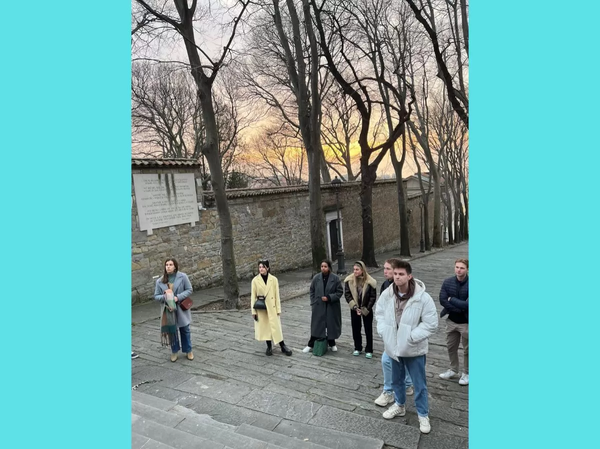 Some of the participants are standing on the steps leading to the San Giusto cathedral. Some leafless trees are behind them. On the background, a beautiful sunset with orange lights.