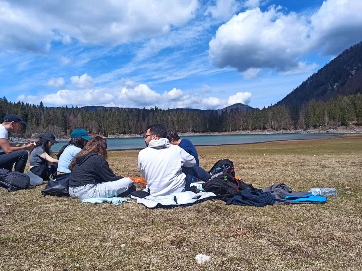 Some of the participants are sitting on the grass with their backs at the camera. We can see the lake in front of them, as well as trees, mountains and a blue sky on the background.