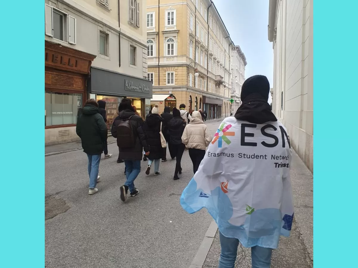 The participants are walking down one of the streets of Trieste with their backs at the camera. On the foreground, one of the ESN volunteers is wearing the section's flag aroung his shoulders.