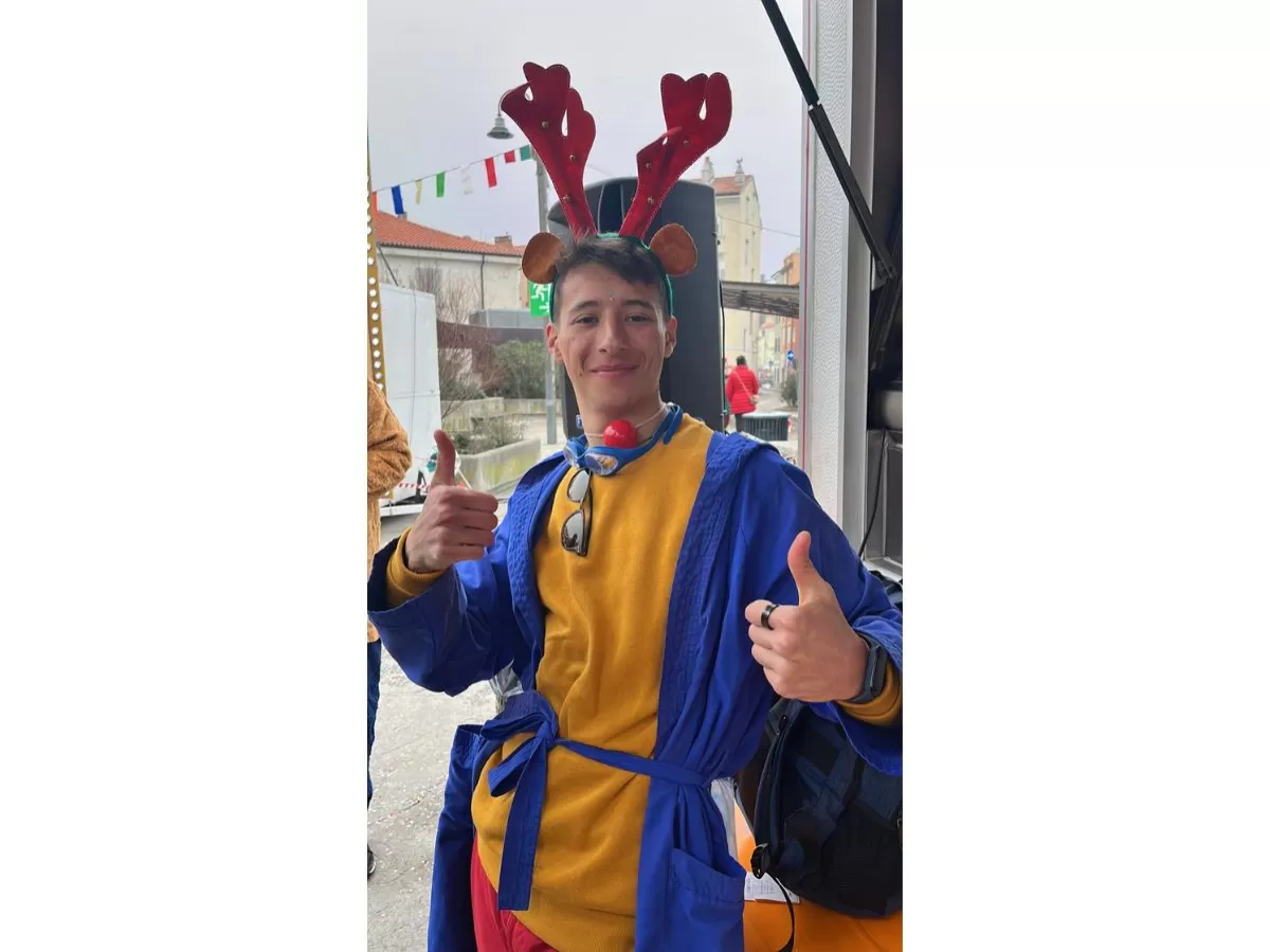 The picture shows on of our voluteers smiling in his carnival costume. He is wearing a yellow sweatshirt with red trousers, a blue bathrobe, a red plastic nose and red reindeer horns.