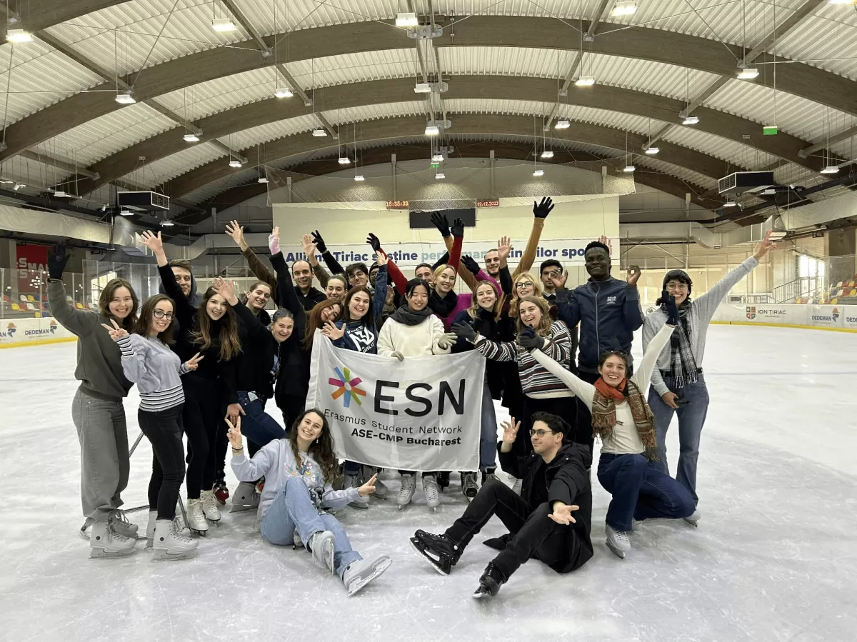A group of smiling people gathered on an indoor ice rink, posing for a photo. Some individuals are standing while others are crouching or kneeling on the ice, and many are waving or making joyful gestures. Everyone is wearing winter clothing and ice skates. In the center, there is a banner with the text "ESN" indicating they may be part of the Erasmus Student Network. The rink is surrounded by barriers and the ceiling structure is visible above, suggesting the setting is a covered sports arena.