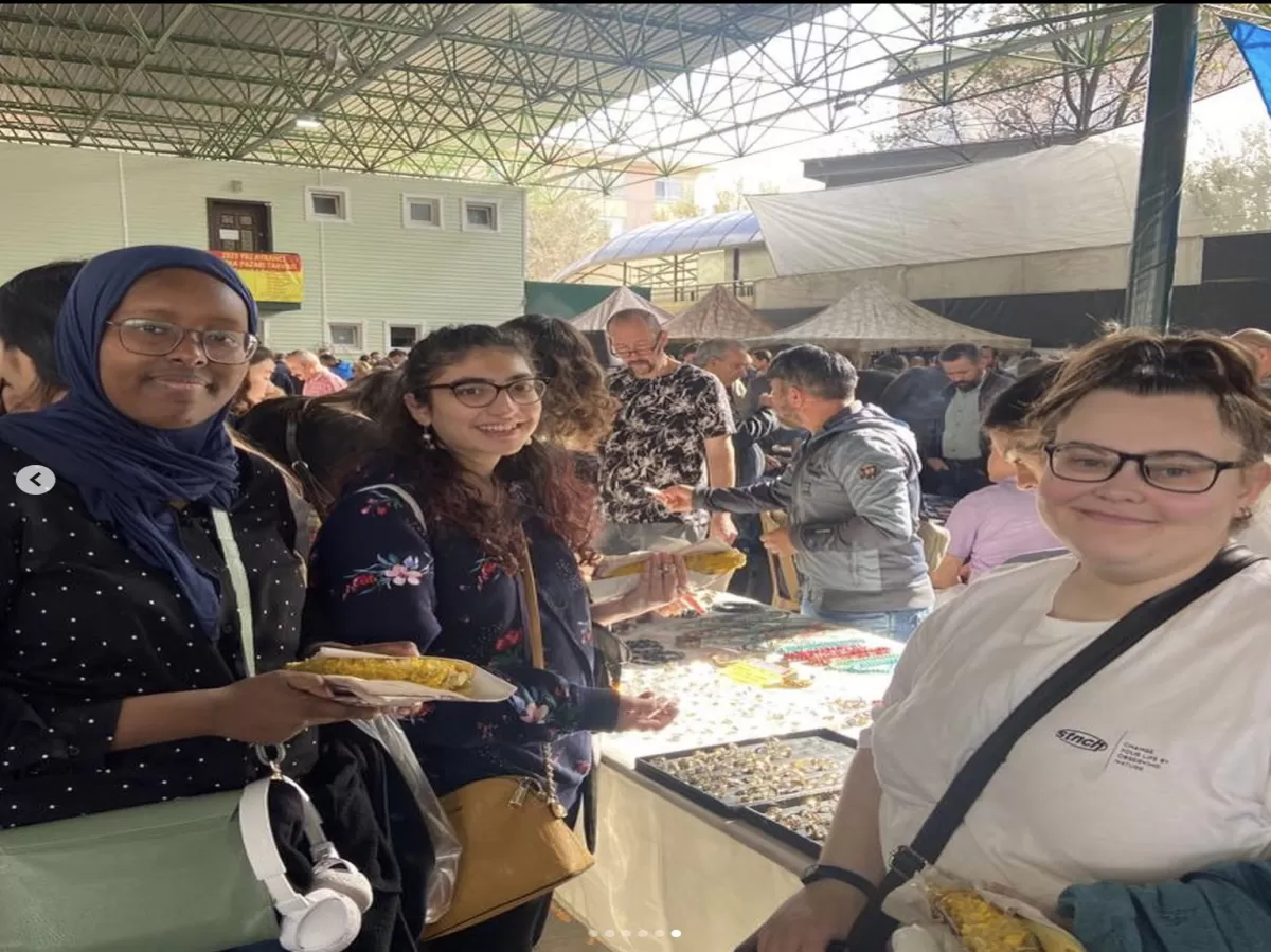 3 Erasmus students posing for the photo in front of a table with jewelry and vendors in the background. They are holding grilled corn cobs in their hands.