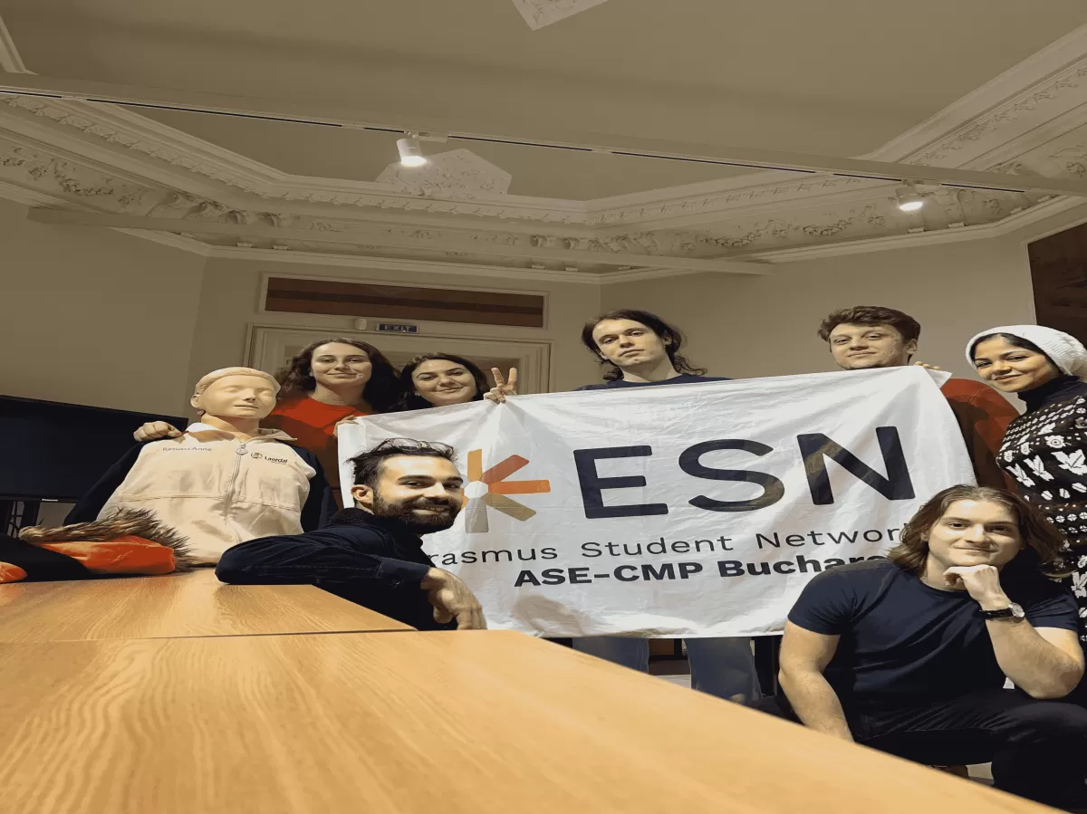 A group photo featuring six participants, the first aid coordinator, and a CPR mannequin, all smiling for the picture. They are holding the ESN (Erasmus Student Network) flag in front of them, conveying a sense of unity and collaboration during a first aid training session.