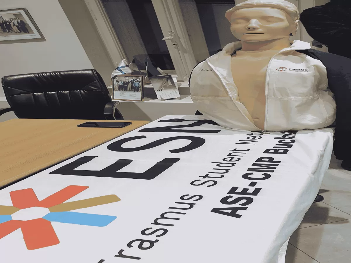 A CPR mannequin positioned alongside the ESN (Erasmus Student Network) flag. The scene illustrates a context related to first aid training, with the mannequin representing a focus on life-saving techniques within the ESN community.