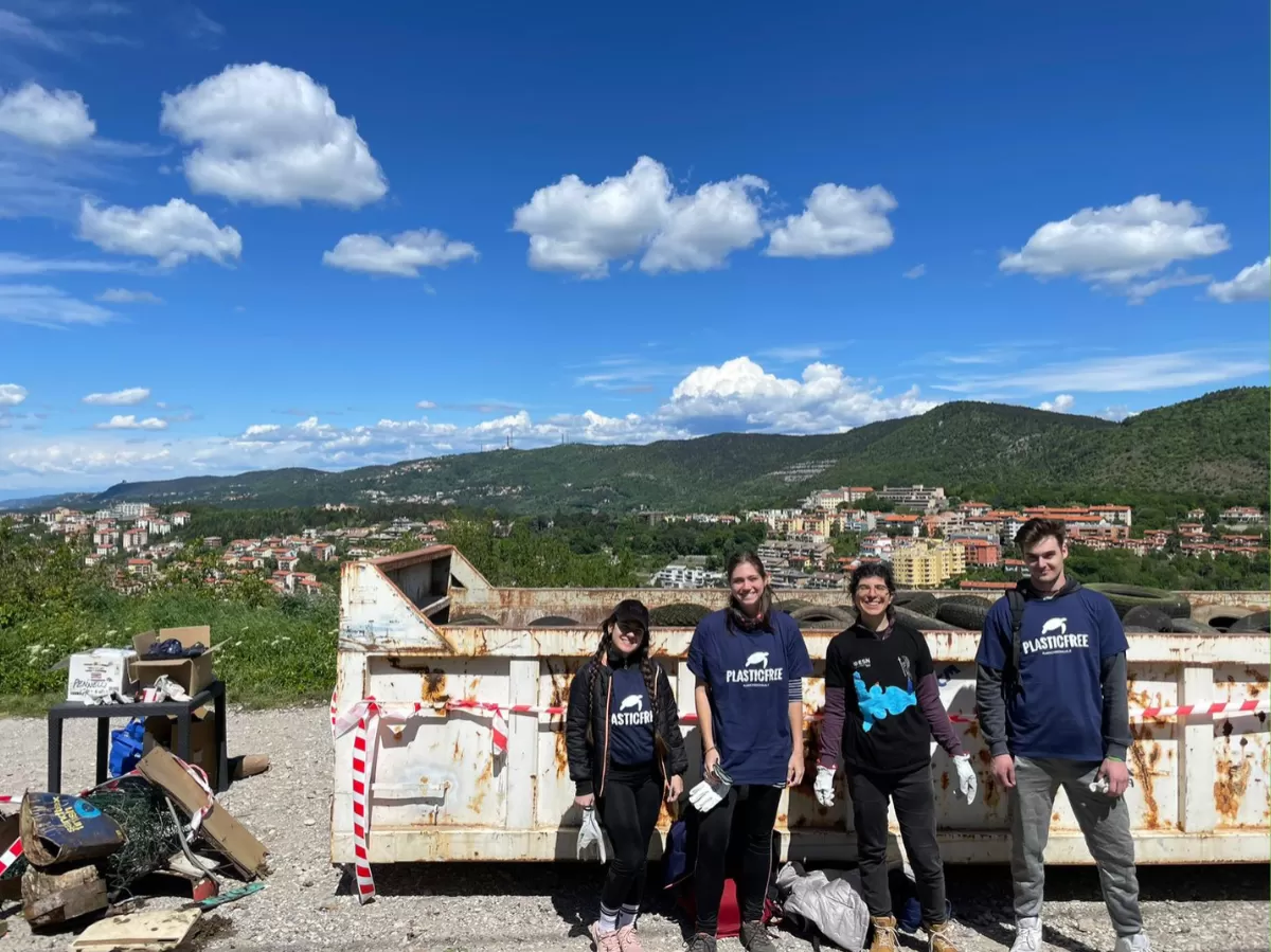 Group picture of the four participants from ESN. On the background there is a blue sky with some clouds and the mountains are visible. The four guys, three girls and a boy, smile at the camera. Three of them wear the Plastic Free t-Shirt and one of them wears the ESN Trieste t-shirt. Behind them stand some of the garbage collected and the huge container full of the tyres disposed of