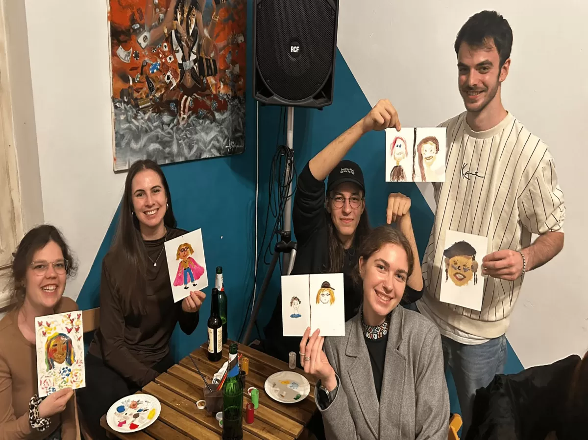 The image shows a group of five people in a cheerful indoor setting, engaged in a painting activity. They are seated around a table with paint supplies, each holding a painting. The atmosphere is friendly and creative, with colorful artwork displayed on the walls in the background. It appears to be a social gathering focused on art and enjoyment.