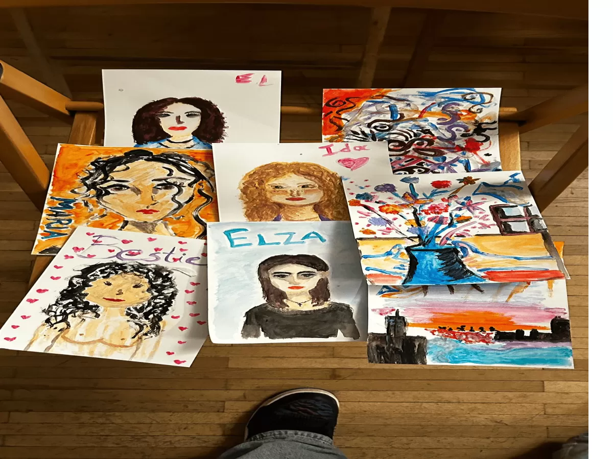 The image shows a collection of colorful paintings on a wooden floor, each created in a different style and depicting various subjects. Some feature portraits with unique styles and names like "Elsa" and "Bestie," while others show scenic or abstract themes. A pair of legs in jeans and a shoe visible at the bottom suggests the viewer's perspective. Overall, the artworks offer a diverse and personal expression of art.