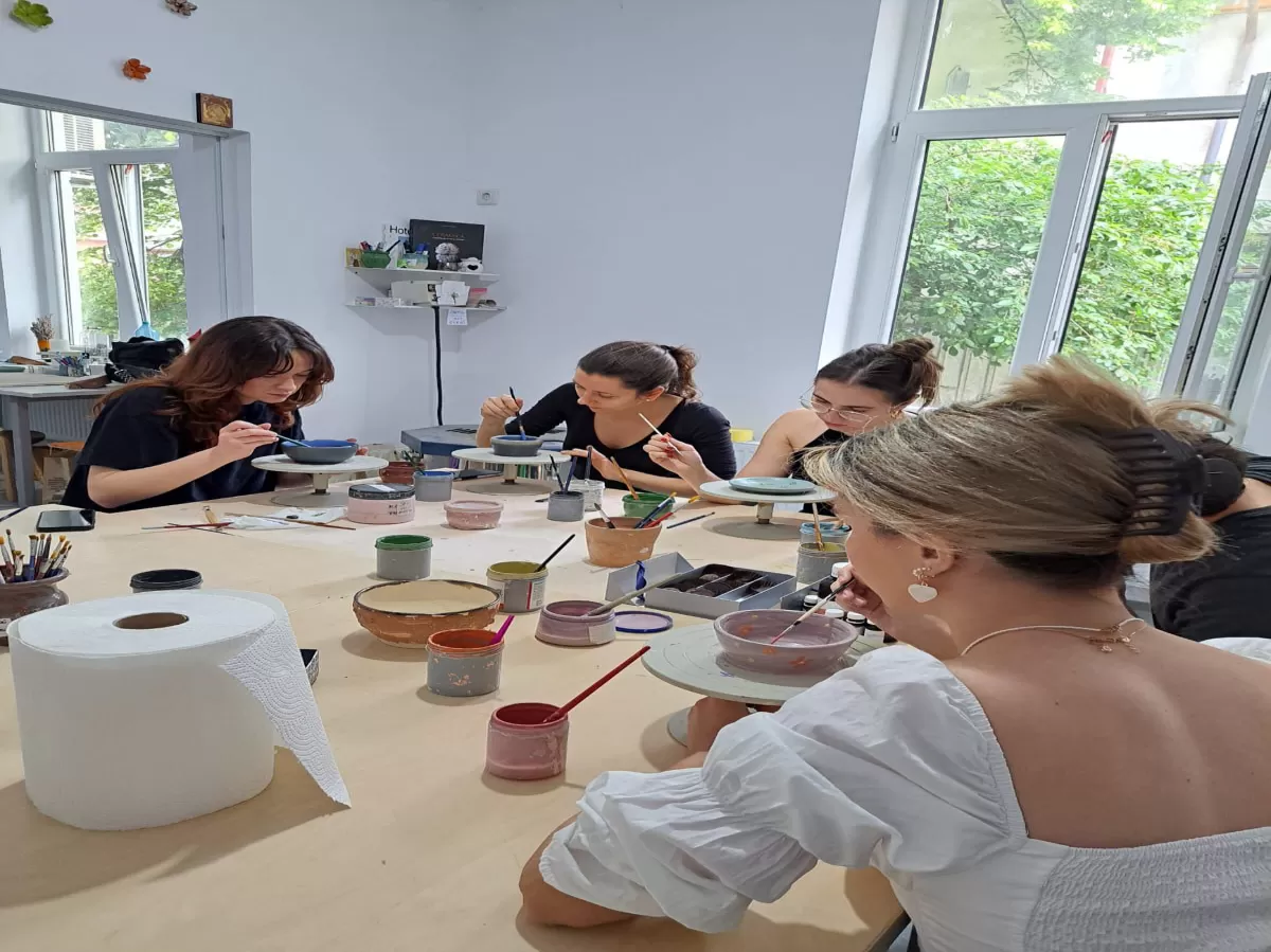 This image captures a group of four women participating in a pottery painting workshop. They are focused on decorating ceramic objects using various colors and brushes. The women are seated around a table in a bright space, likely an art studio, with large windows letting in natural light. On the table, there are various materials needed for pottery painting, including containers of paint and rolls of absorbent paper. The atmosphere seems relaxed and creative.