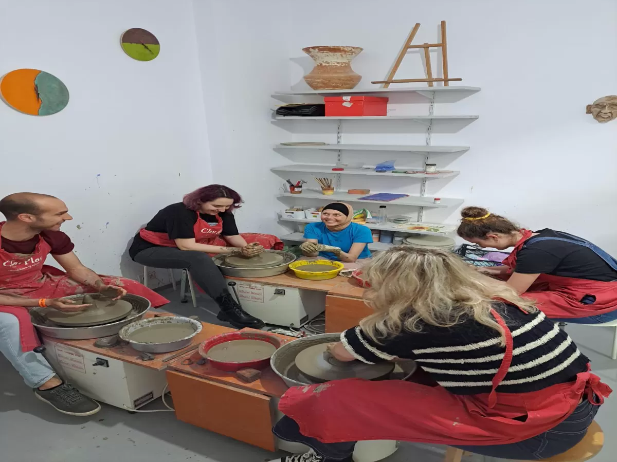 The image captures a group of five people engaged in pottery making in a ceramic workshop. The participants are focused on shaping the clay on potter's wheels, wearing aprons for protection. Various ceramic objects are visible on shelves, including colorful vases and plates, adding a decorative and artistic background to the scene. The atmosphere is creative and interactive.