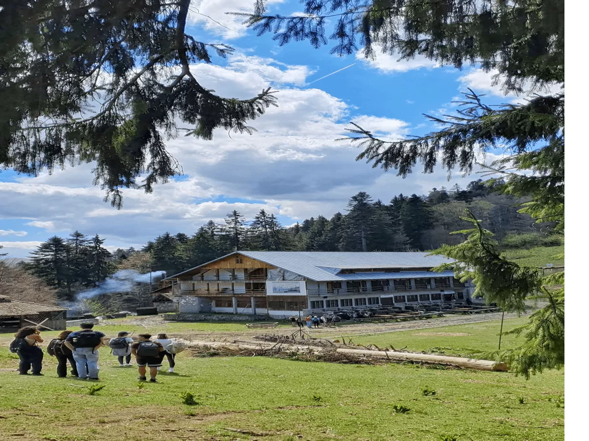 The image depicts a group of people walking towards a traditional mountain cabin in a scenic natural setting. Surrounded by lush forest and greenery, the cabin exudes a cozy vibe with smoke rising from the chimney. The serene atmosphere and picturesque landscape make it an ideal escape for outdoor activities or a peaceful retreat.