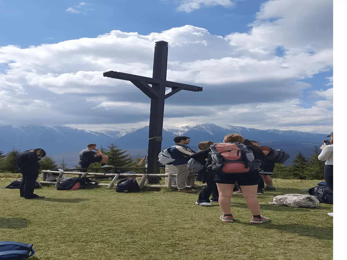 The image shows a group of people on a mountain hike. In the center, there is a large wooden cross on a mountain peak, indicating a possible pilgrimage site or important landmark for hikers. Behind the cross, there are stunning views of distant mountain ranges under a partially cloudy sky. The hiking group consists of men and women of varying ages, dressed in hiking gear. Some are standing and chatting, while others are seated on a bench or on the ground, resting and enjoying the scenery.