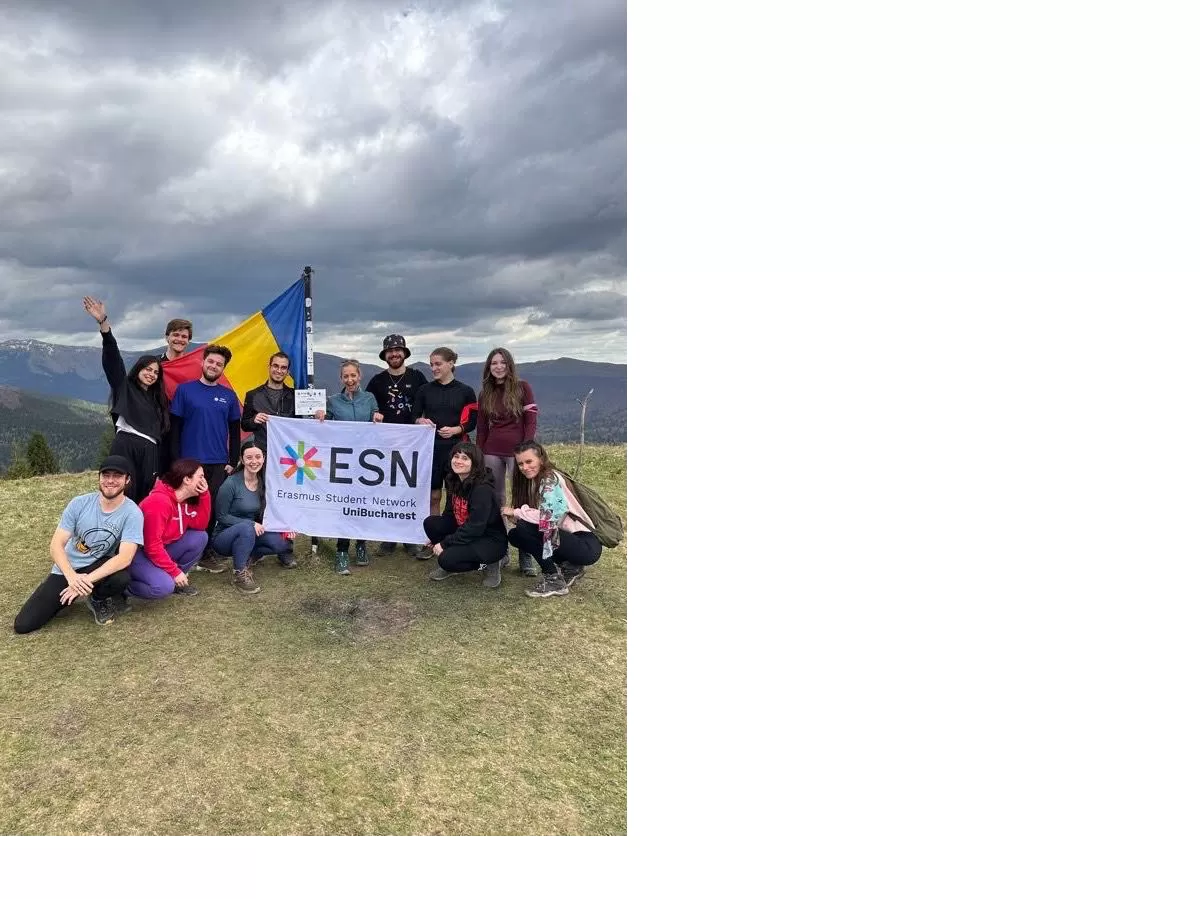 The image depicts a group of fourteen young people sitting on a hill with a mountain backdrop. They hold a large flag with "ESN" and "Erasmus Student Network UniBucharest" on it, along with the Romanian flag. The group looks happy, showing unity during an event likely organized for international students through the Erasmus network.