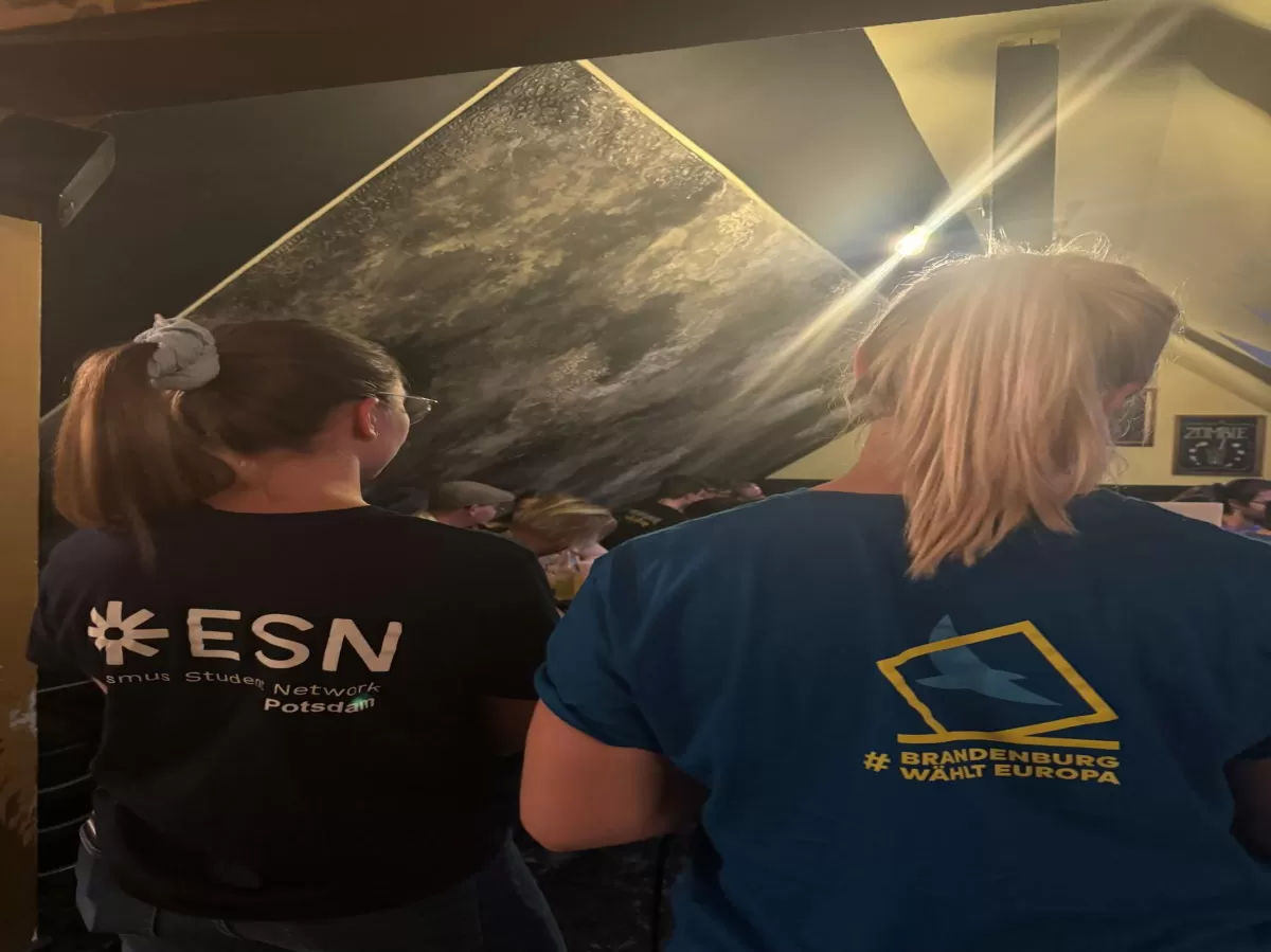 Two girls standing in a bar with their backs towards the camera. The left gils is wearing a shirt with the ESN Potsdam logo and the other girl a shirt from a campaign called "Brandenburg wählt Europa"