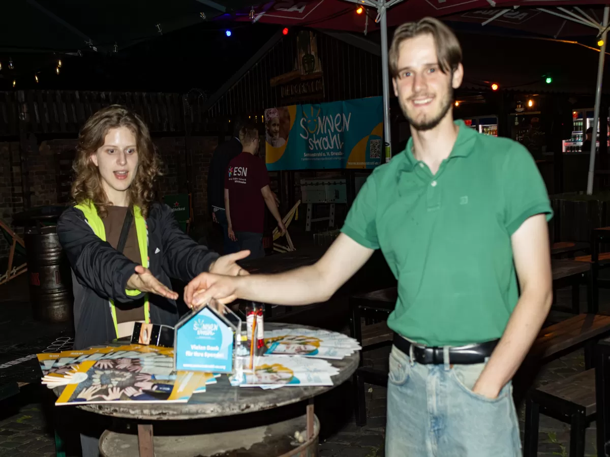 2 persons at a round table with flyers and magazines about Sonnenstrahl e.v. one person is putting money in the donation box