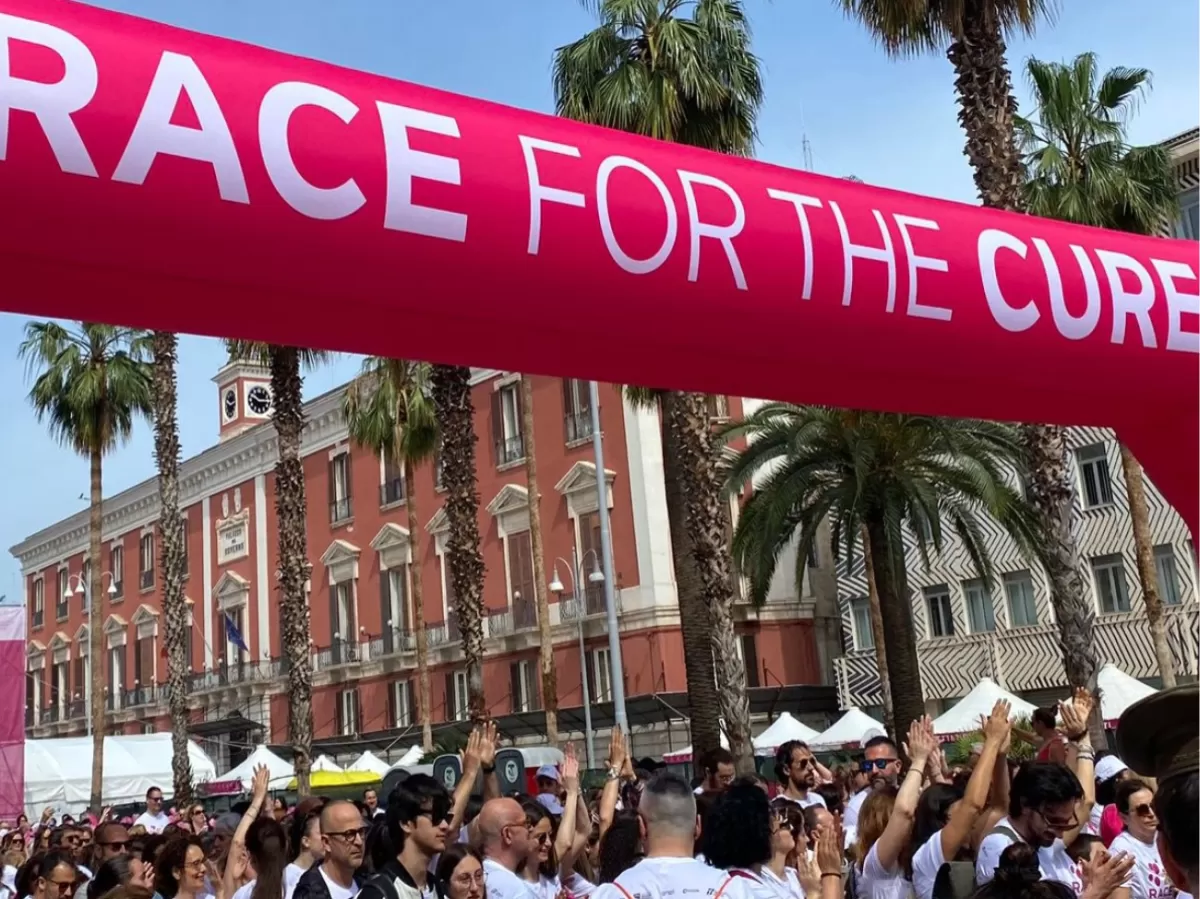 A large pink arch marked “Race for the Cure” greets an energetic crowd. People raise their hands, ready to support the cause, while historic buildings and palm trees can be seen in the background.