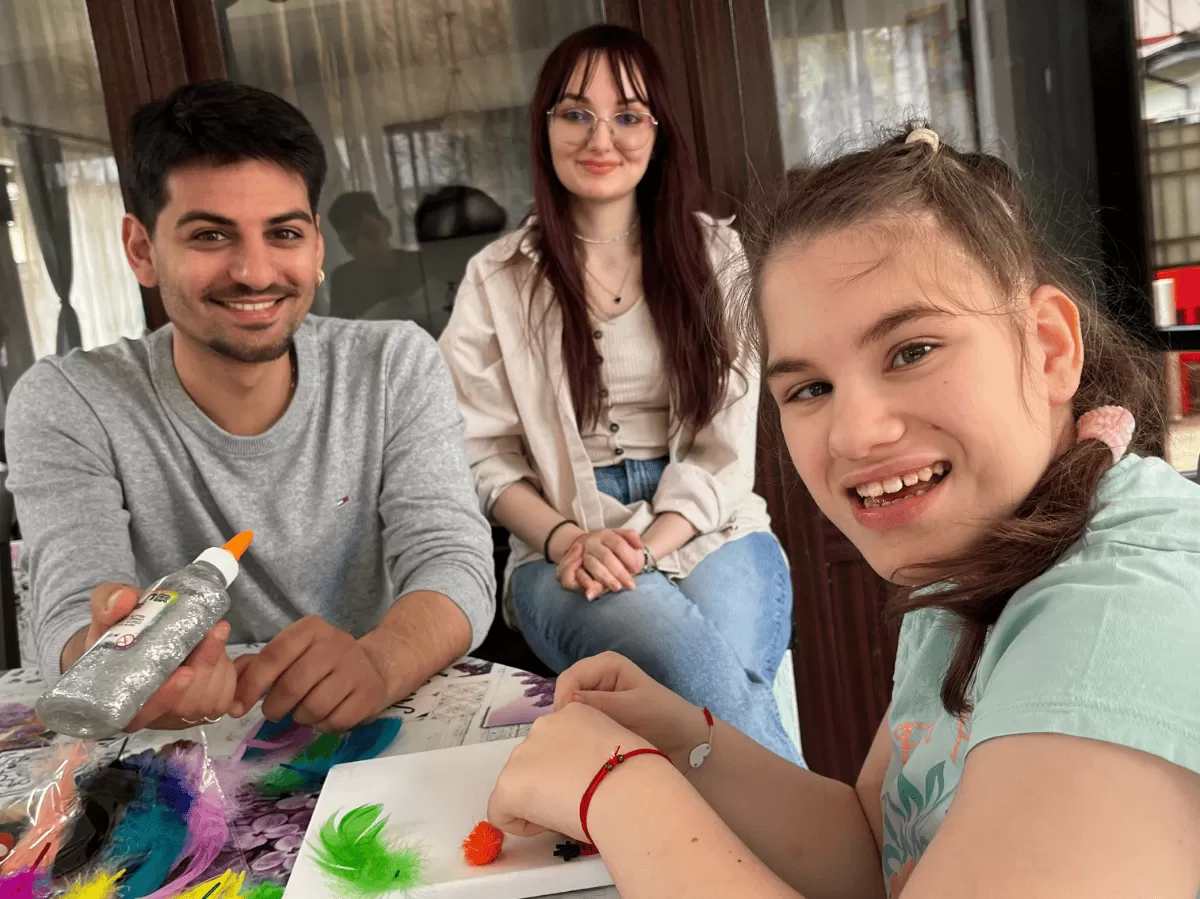A young girl with a neurological condition smiles brightly while engaging in a handcraft activity using colorful feathers and pom-poms. Next to her, a smiling male volunteer holds a bottle of glitter glue, and a female volunteer sits in the background with a gentle smile. The environment is warm and supportive, reflecting a creative and inclusive workshop setting where children receive individualized attention and encouragement.