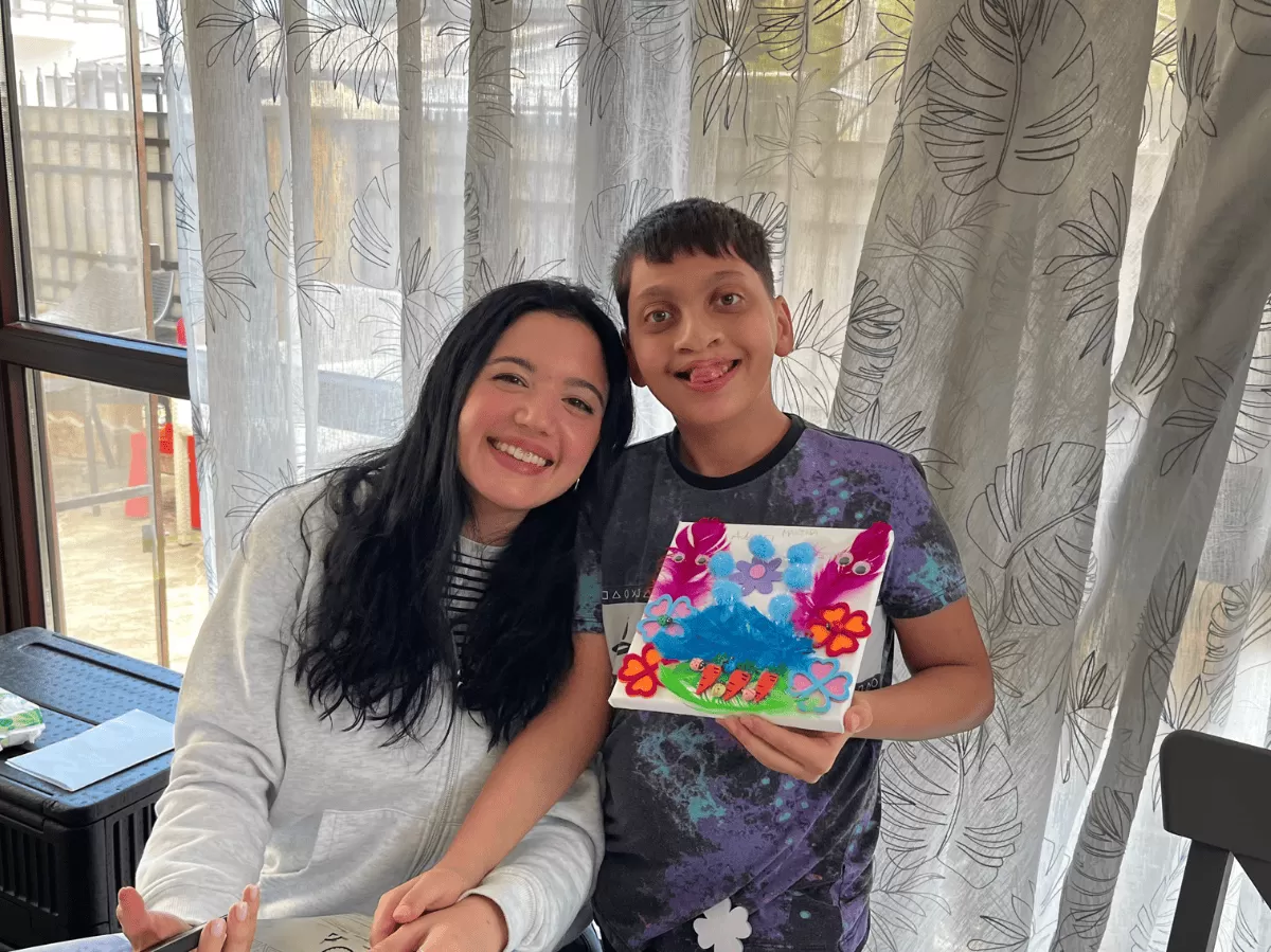 A smiling volunteer sits closely beside a boy with neurological conditions, both beaming with joy. The boy proudly holds up a colorful handmade artwork decorated with bright feathers, foam flowers, and drawings of carrots, created during the workshop. The warm interaction and creative expression highlight the supportive, inclusive atmosphere of the activity.