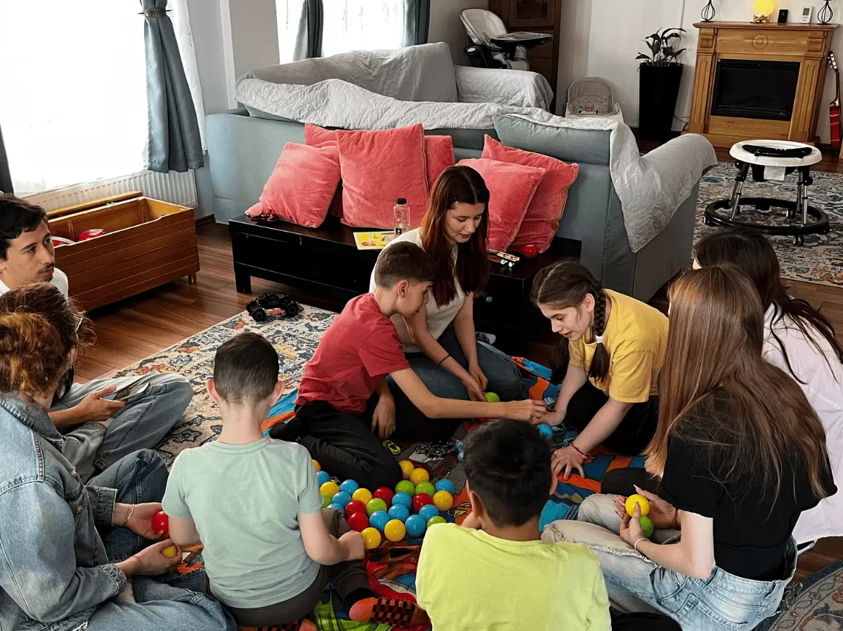 Group of children and adults sitting on the floor, playing with colorful plastic balls in a cozy living room.