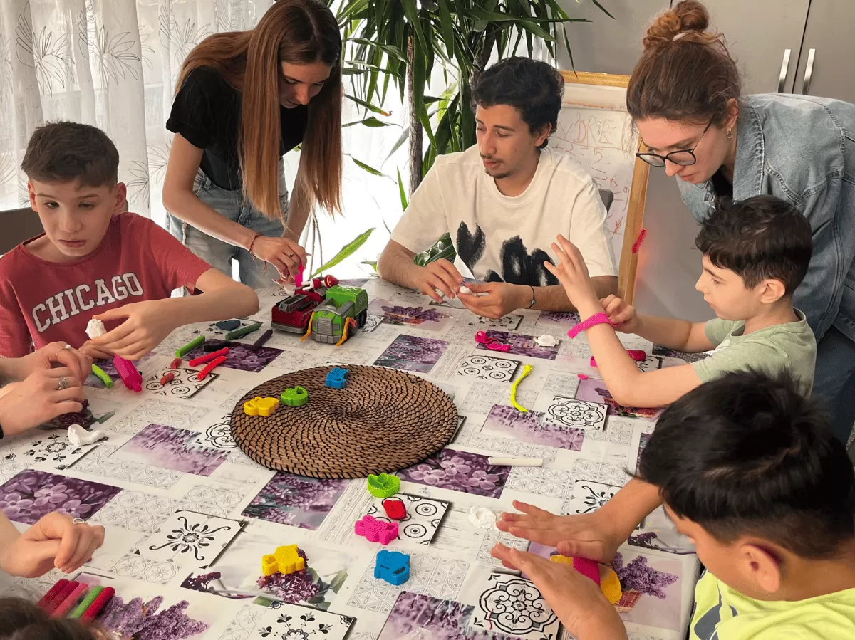 Children and adults sitting around a table, playing with colorful modeling clay and toys in a bright indoor setting.
