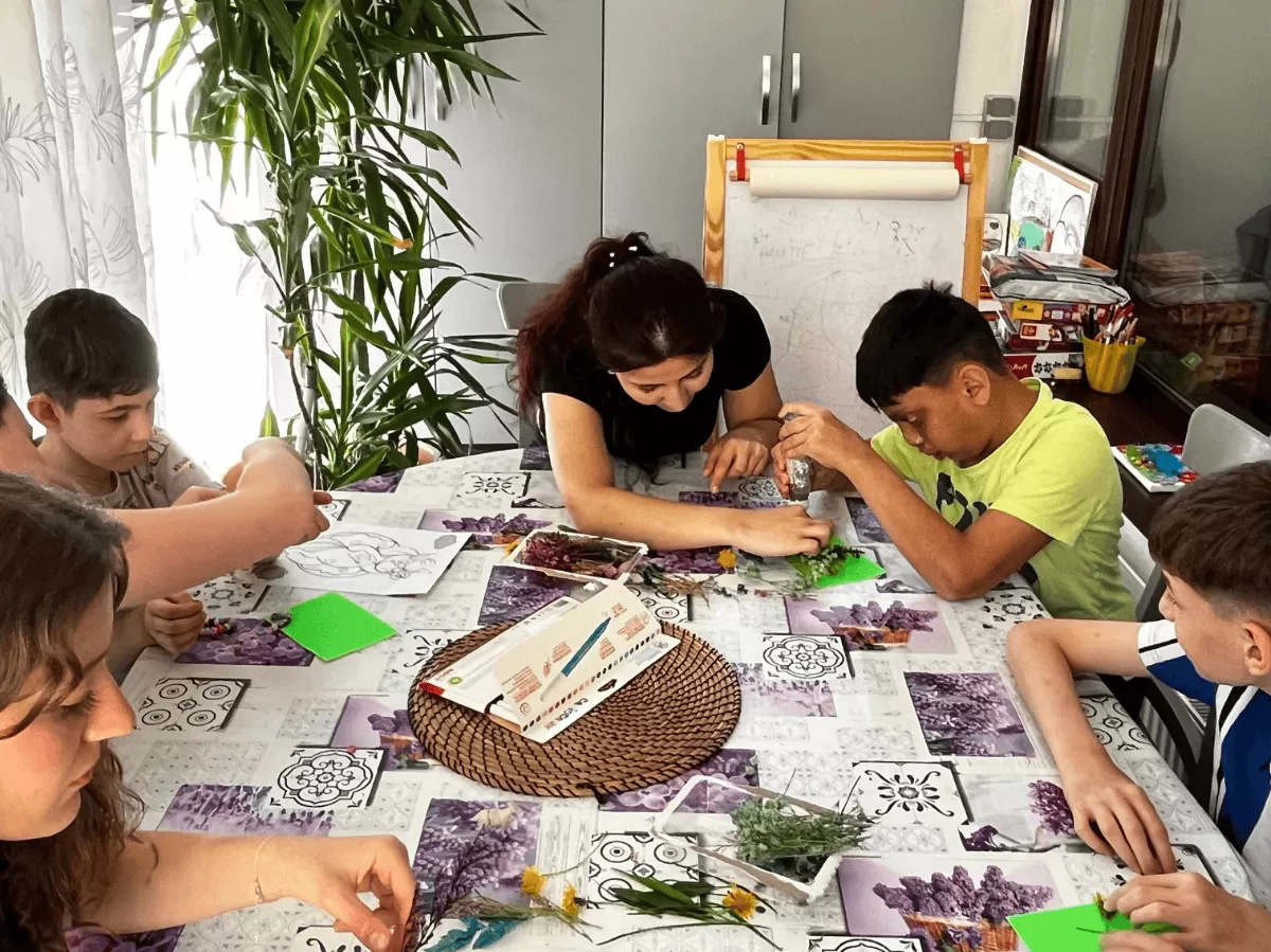 Children and adults sitting around a table, engaging in a crafting activity with natural elements like flowers and leaves, in a bright room with a large plant nearby.