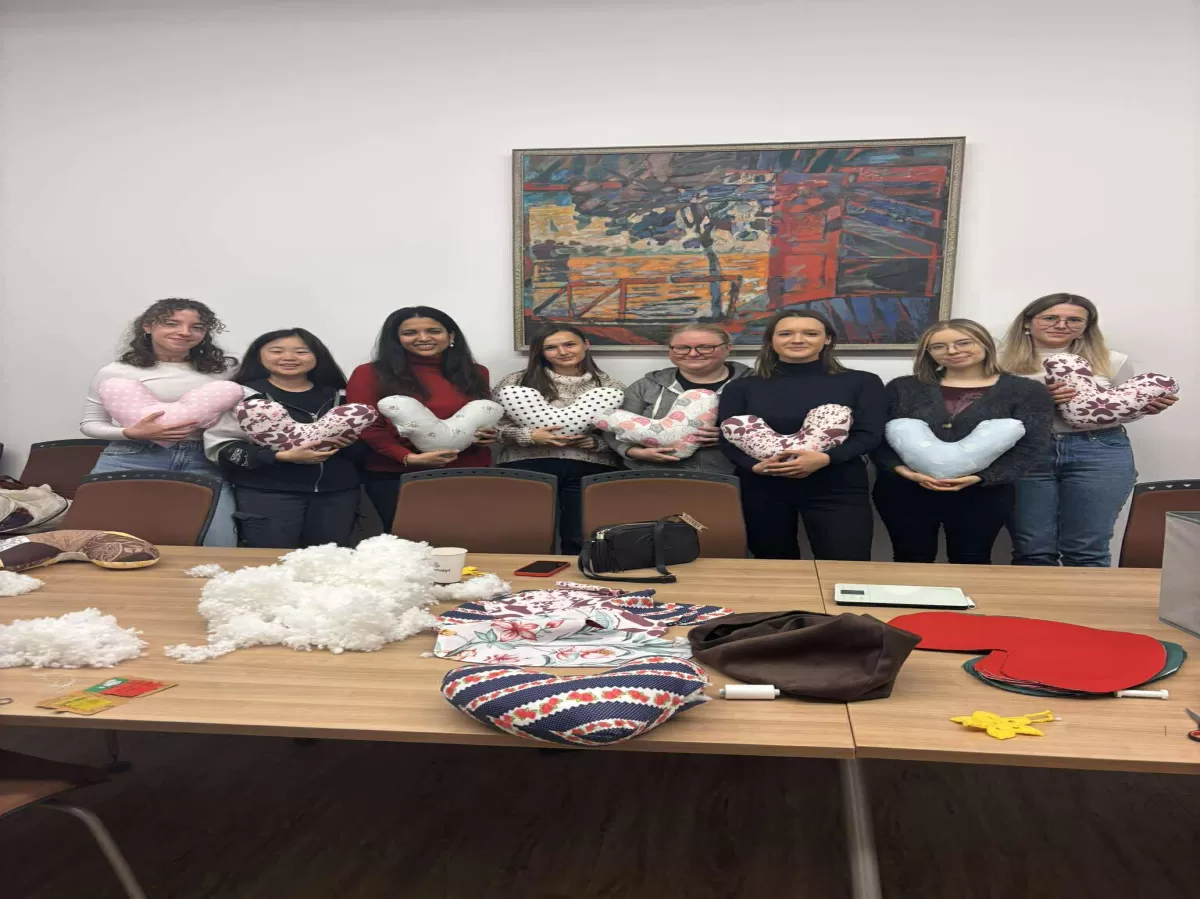 A group of seven women are sitting around a table in a room, each holding a colorful, U-shaped or crescent-shaped pillow with various patterns, such as polka dots and floral designs . In the foreground, the table is covered with crafting materials, including a pile of white stuffing, fabric swatches, and other sewing supplies. A large, abstract painting is hanging on the wall in the background. The scene appears to be a crafting workshop or a similar meeting.
