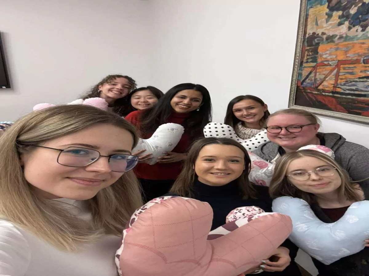 A group of eight smiling women holding creatively shaped pillows, some pink and lung-shaped, others patterned, while posing indoors for a photo.