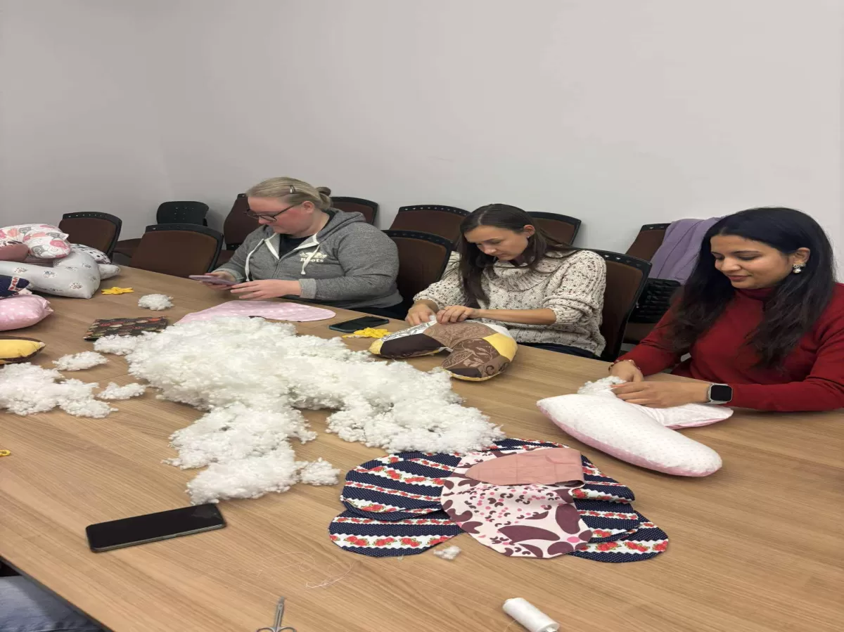 Three women are sitting at a large wooden table working on a craft project, likely making pillows or soft toys. The table is covered with a large pile of white stuffing material, various fabric pieces (some patterned with flowers or geometric shapes), scissors, thread, and mobile phones. The woman on the left wears a grey hoodie, the middle woman wears a patterned sweater, and the woman on the right wears a red top. They are all focused on sewing their respective fabric items.