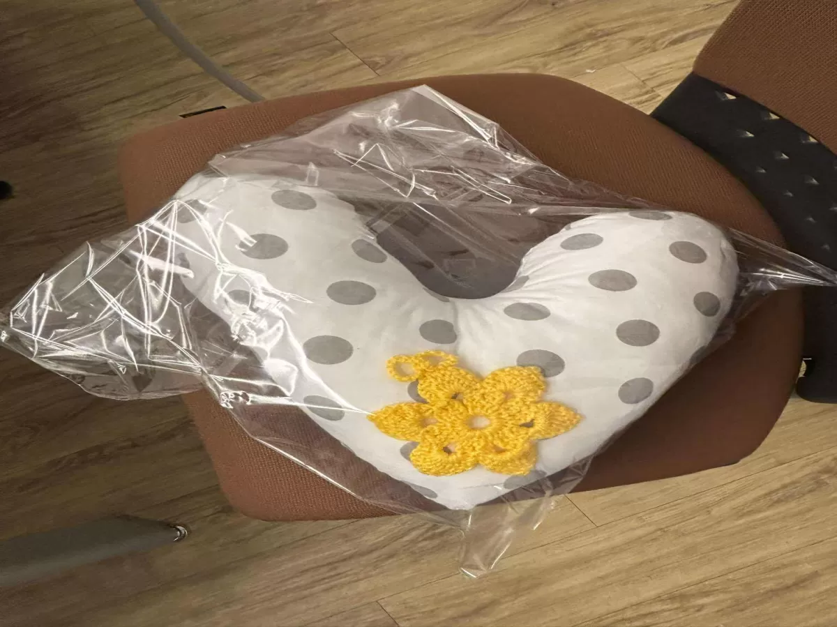 A white, heart-shaped pillow with gray polka dots and a yellow crocheted flower decoration, wrapped in clear plastic packaging and sitting on a brown chair.